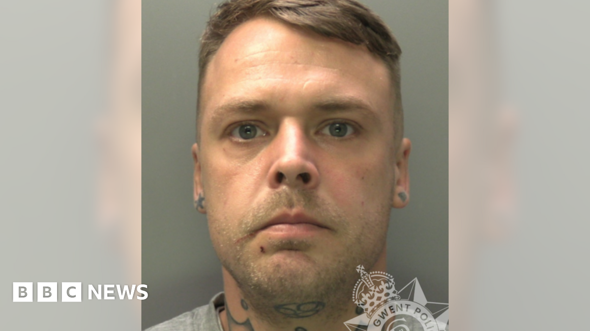 Custody photo of Kai Pennell. He has short, light brown hair with shaved sides, blue eyes and stubble. He has tattoos on his neck and earlobes.