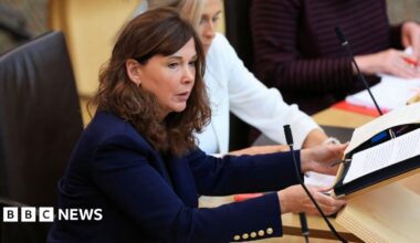 Peter Murrell looking off to his right while sitting in a room. He is wearing a black suit, white shirt and dark coloured tie.