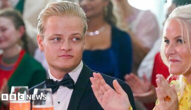 A young man with blond hair and a bow tie looks on as a woman in a yellow dress claps