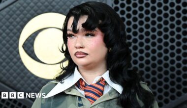 Lola Young has long black hair, she is wearing a dark green jumper with a large white collar over the top, she has a orange and blue striped tie on. She is holding a GRAMMY award.