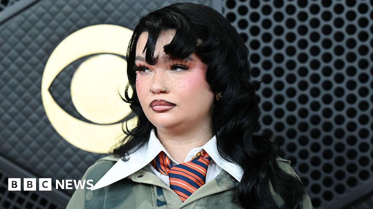 Lola Young has long black hair, she is wearing a dark green jumper with a large white collar over the top, she has a orange and blue striped tie on. She is holding a GRAMMY award.