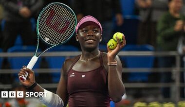 Victoria Mboko plays a backhand. She has black hair, tied back, and is wearing a pink cap and maroon dress. Her right arm is bandaged.