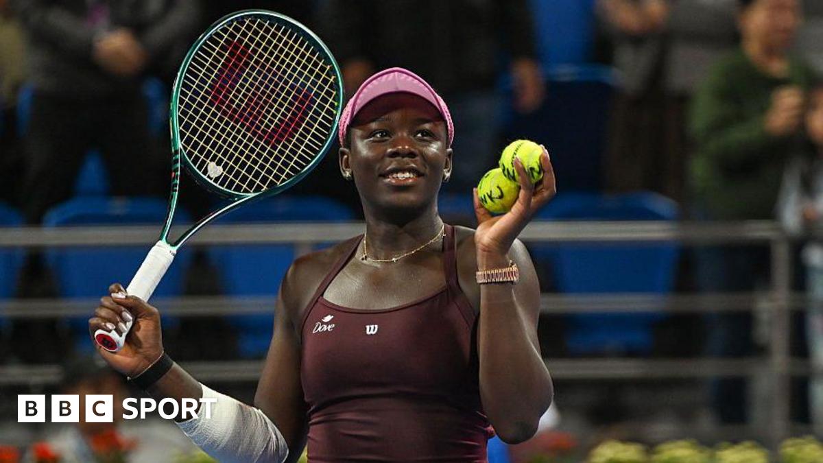 Victoria Mboko plays a backhand. She has black hair, tied back, and is wearing a pink cap and maroon dress. Her right arm is bandaged.