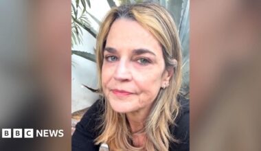 A woman wearing a dark top and jeans stands in quiet contemplation by a flower memorial outside Nancy Guthrie's home