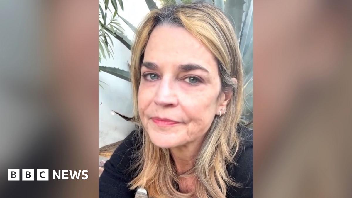 A woman wearing a dark top and jeans stands in quiet contemplation by a flower memorial outside Nancy Guthrie's home