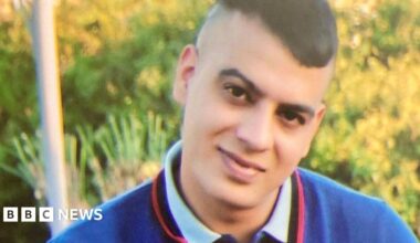 Choudry sitting on an outdoor bench, wearing a short‑sleeved navy blue polo shirt with red and white trim on the collar and sleeves. The shirt has a circular white emblem on the left side of the chest. He is also wearing blue jeans and a wristband on their right wrist.
