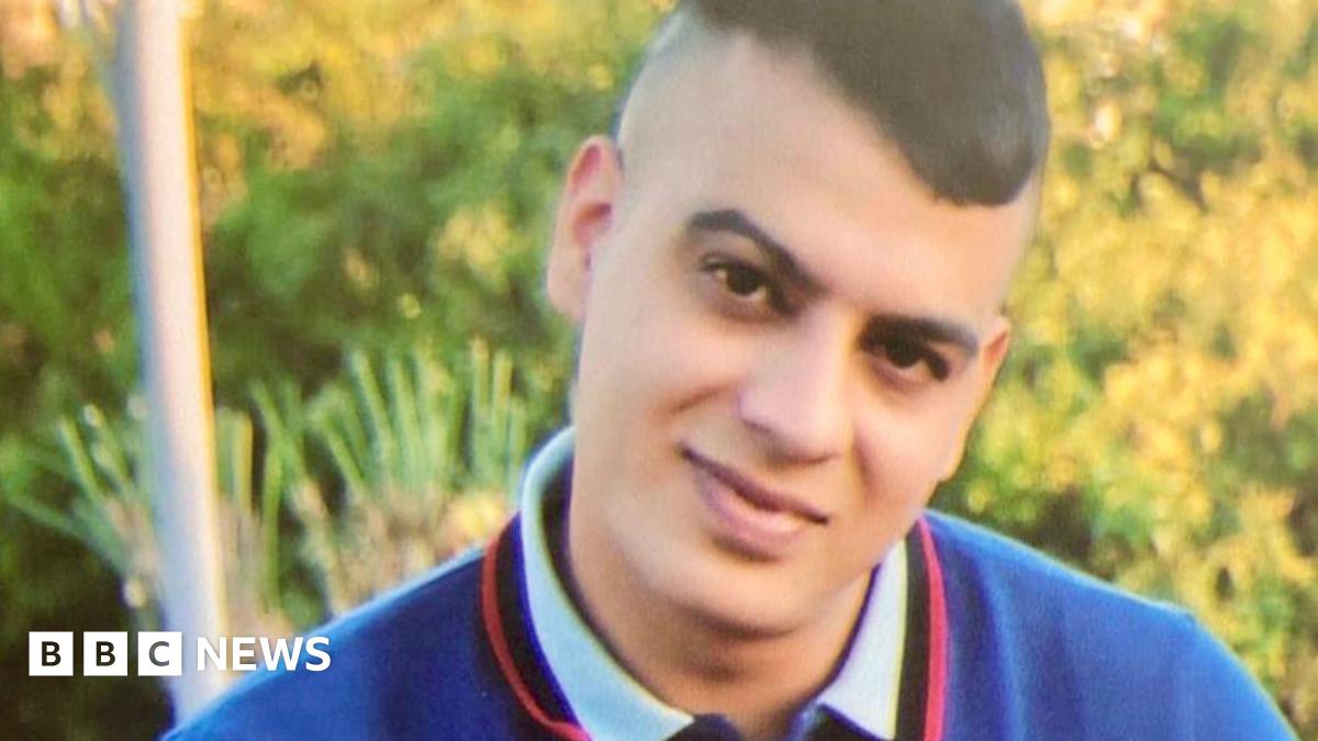 Choudry sitting on an outdoor bench, wearing a short‑sleeved navy blue polo shirt with red and white trim on the collar and sleeves. The shirt has a circular white emblem on the left side of the chest. He is also wearing blue jeans and a wristband on their right wrist.
