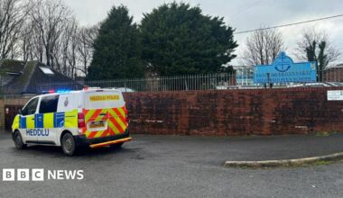 A police van and car are outside the entrance to the school. A sign with the school name can also be seen.