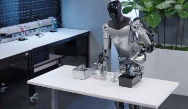 Tesla’s Optimus humanoid robot works at a lab table during a demo, showing AI-driven hands handling small parts.