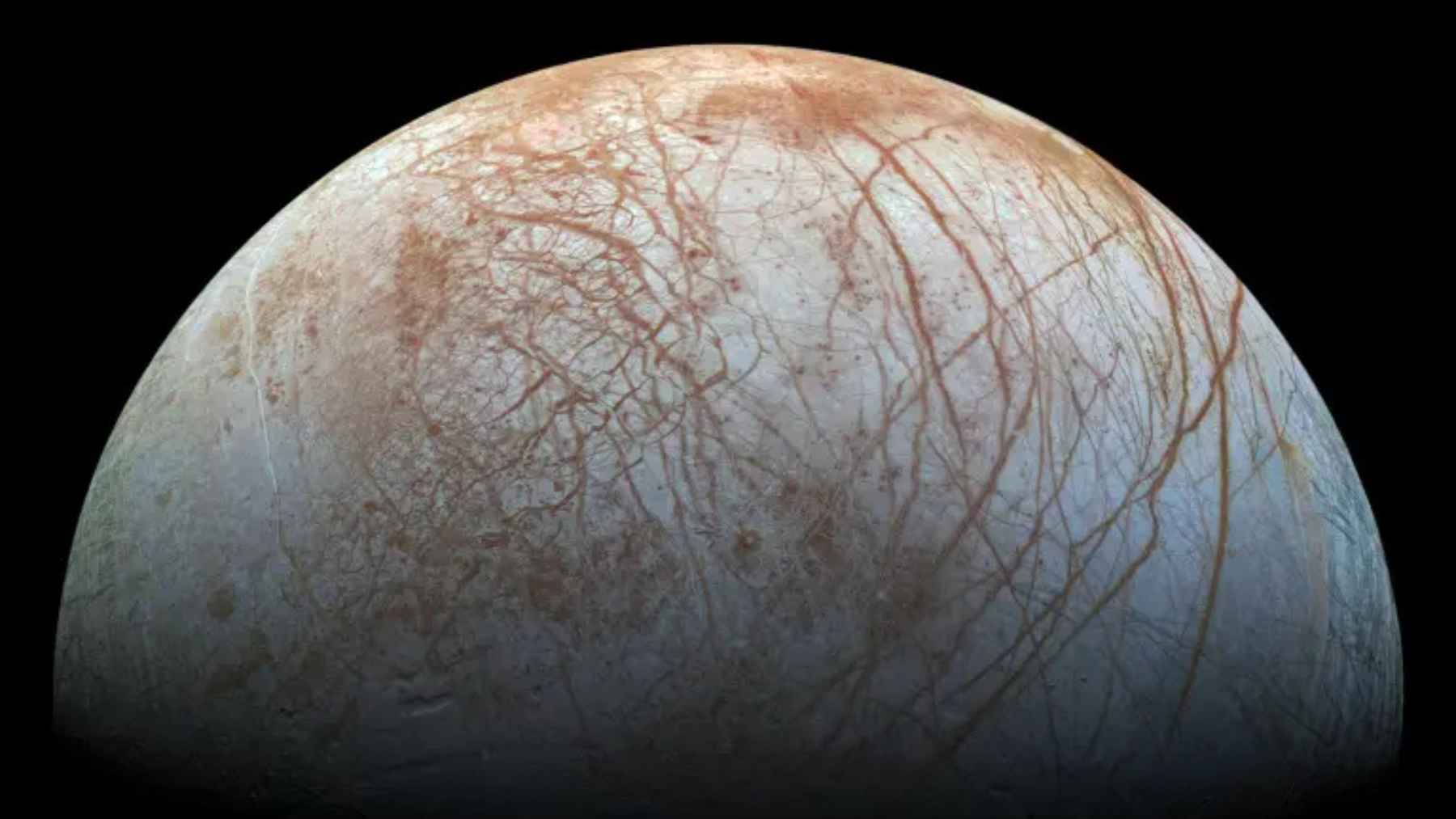 Europa, Jupiter’s icy ocean moon, showing a bright cracked surface with reddish-brown streaks against black space.