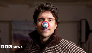 Radio 1 DJ Greg James stood behind a black tandem bike with his left hand on the handlebars and his right arm in the arm with his fish clenched. He is wearing blue trousers and a red jacket and has a red nose on his nose, with a white cap. The photo is taken in a studio setting so there are two big bright photography lights behind him and a canvas style picture of Edinburgh Castle.