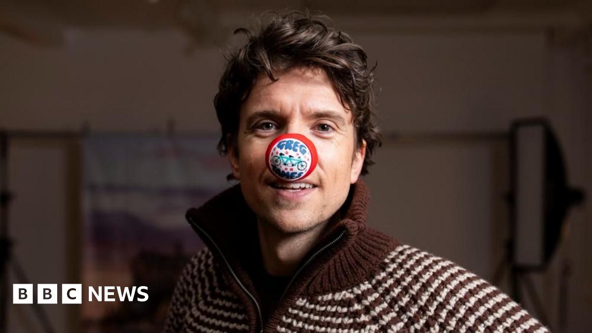Radio 1 DJ Greg James stood behind a black tandem bike with his left hand on the handlebars and his right arm in the arm with his fish clenched. He is wearing blue trousers and a red jacket and has a red nose on his nose, with a white cap. The photo is taken in a studio setting so there are two big bright photography lights behind him and a canvas style picture of Edinburgh Castle.
