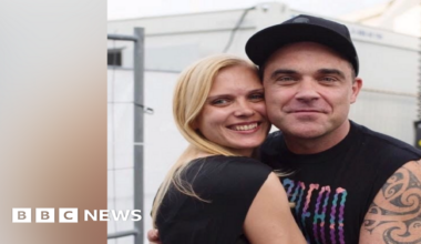 A woman with blonde hair is hugging Robbie Williams, who has his arms around her. They both smile at the camera.