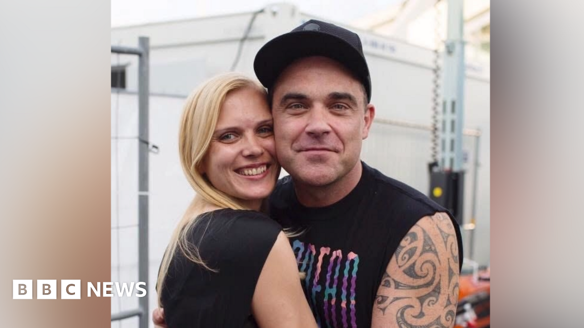 A woman with blonde hair is hugging Robbie Williams, who has his arms around her. They both smile at the camera.