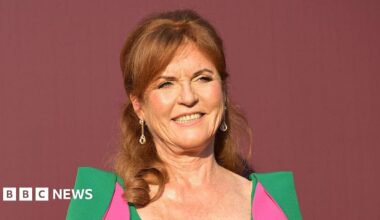 Sarah Ferguson stood in front of a dark red back drop. She is wearing a green dress with pink parts to it.