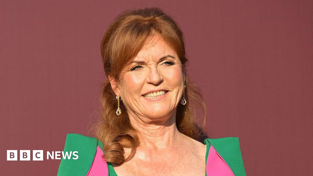 Sarah Ferguson stood in front of a dark red back drop. She is wearing a green dress with pink parts to it.