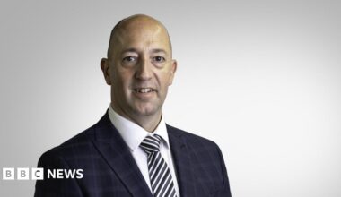 Paul Vane pictured in a black check suit. Black and white stripey tie and white shirt. He is smiling. He is middle aged with a bald head. He's being photographed in a studio, with a blank background.