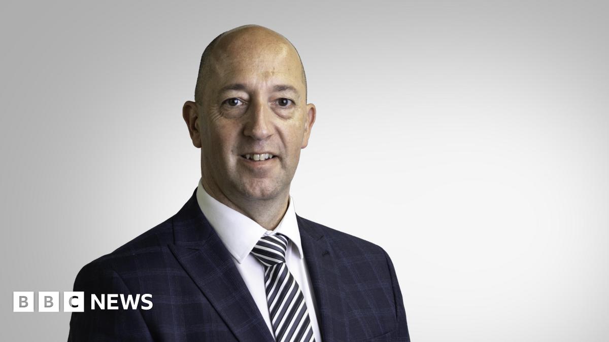Paul Vane pictured in a black check suit. Black and white stripey tie and white shirt. He is smiling. He is middle aged with a bald head. He's being photographed in a studio, with a blank background.