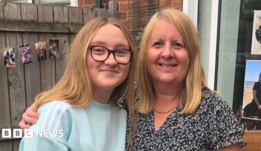 A daughter and mother standing arm in arm in a back garden, with a wooden fence, patio door and various colourful photos in the window and hanging from the fence. The daughter, a young woman, has long strawberry blond hair and wears purple-framed glasses and a light blue jumper with white stripes on the shoulder. The mother has similar hair and wears a black floral blouse, with a thin gold necklace around her neck.