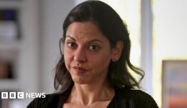 A woman with dark medium length hair and brown eyes is looking at the camera. She is wearing a black cardigan and a chain with a small pendant around her neck. The background is blurred, but shows a bookcase and light coming through a window.