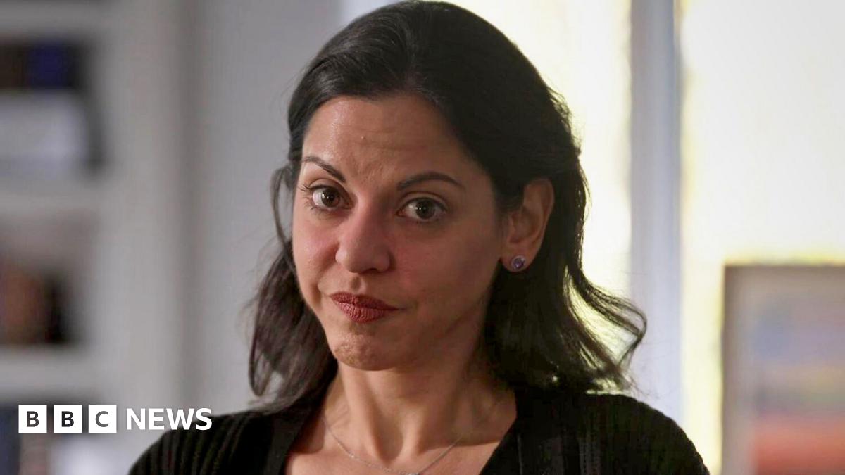 A woman with dark medium length hair and brown eyes is looking at the camera. She is wearing a black cardigan and a chain with a small pendant around her neck. The background is blurred, but shows a bookcase and light coming through a window.