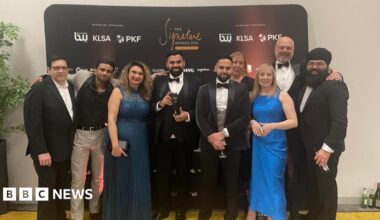 Nine people are standing together in front of a black banner with gold writing on it which reads "the signature awards 2026". There are six men and one woman. The men are dresses in suits and ties, and the women are in dresses. In the centre is Manny Athwal, who is in a black suit with a white shirt and black dickie bow tie. He is holding a black trophy.