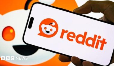 Reddit logo displayed on a smartphone held by hand, with a larger version of the company's logo behind it.