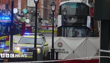 A white double decker bus with the front window smashed, parked behind a white Dublin Fire Brigade sheet. There is a fire engine and a garda car parked beside it.
