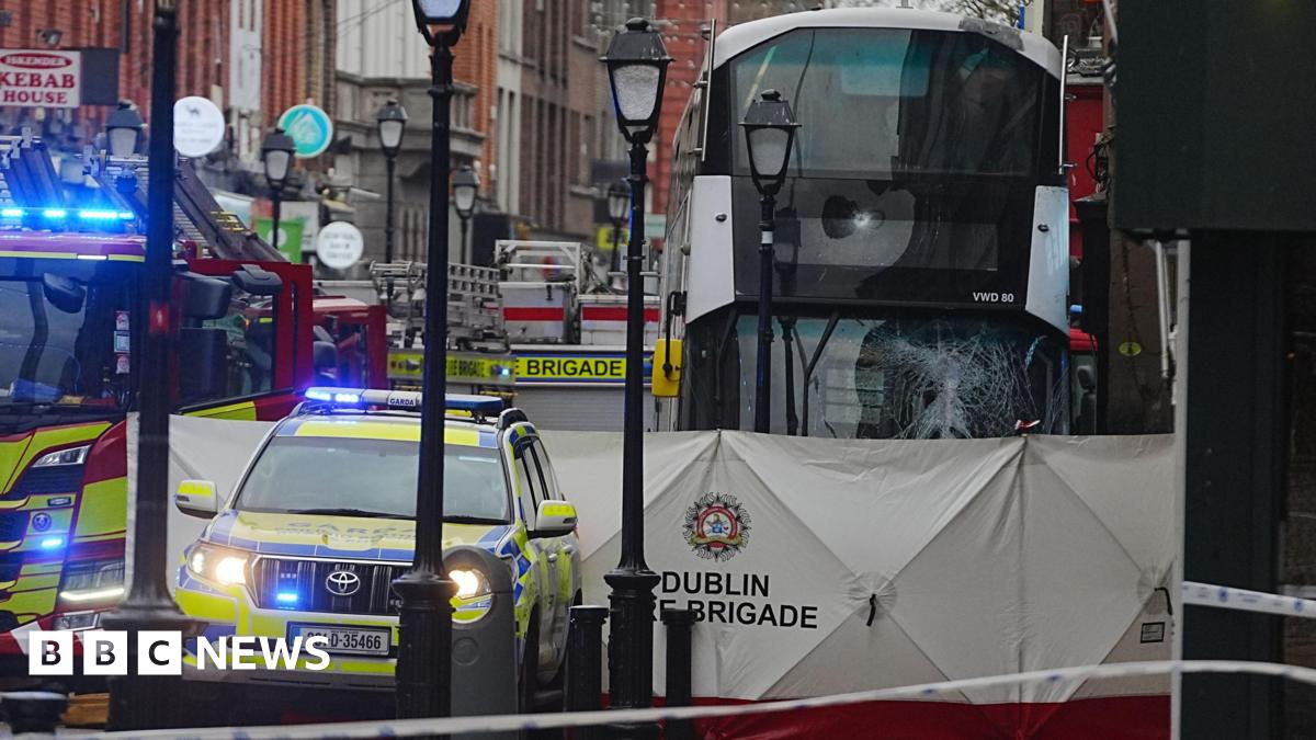 A white double decker bus with the front window smashed, parked behind a white Dublin Fire Brigade sheet. There is a fire engine and a garda car parked beside it.