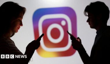 Two teenagers stand in front of a large logo of Instagram. Both are on their phones.