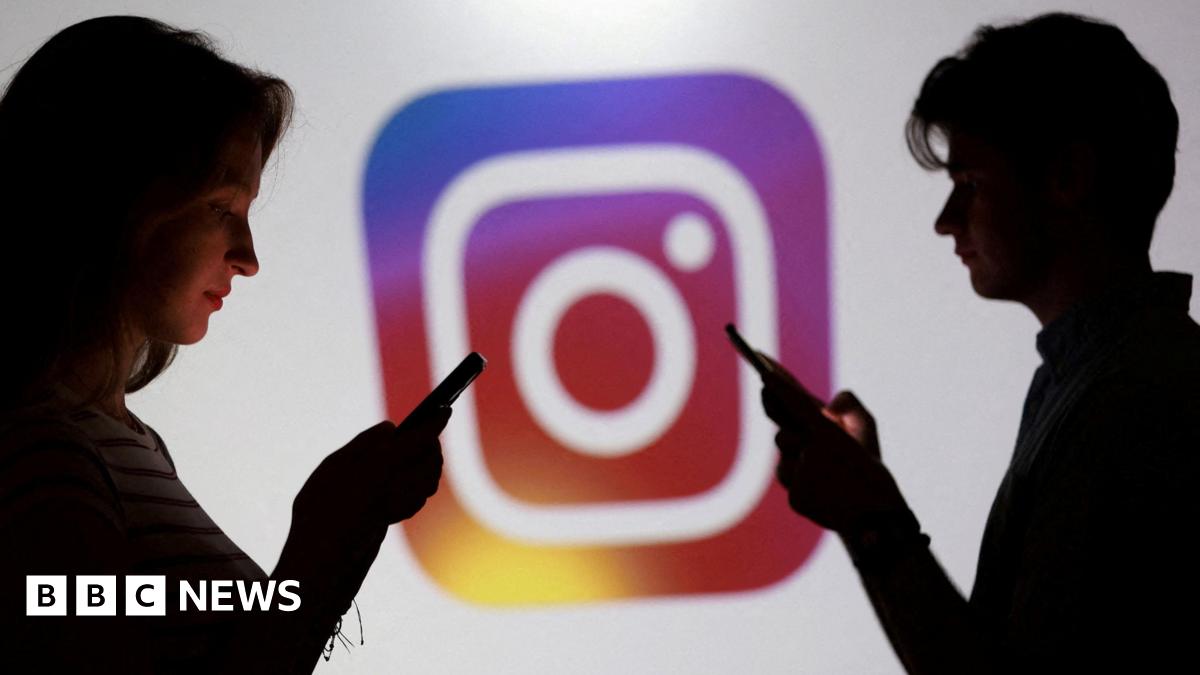 Two teenagers stand in front of a large logo of Instagram. Both are on their phones.