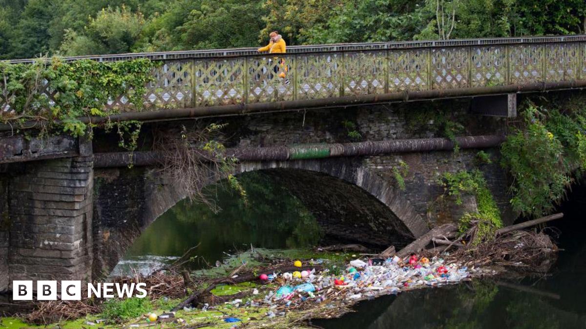 Plastic bottles collect in a river under a bridge, with a walker in an orange coat crossing the bridge above.