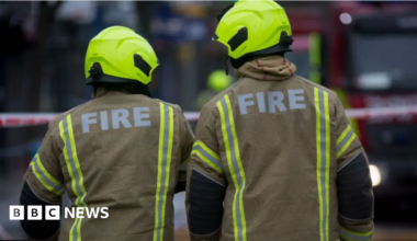 The back of two fire fighters. They are wearing a beige oversized jacket with the word "fire" printed in bold white lettering and wear illuminous yellow helmets