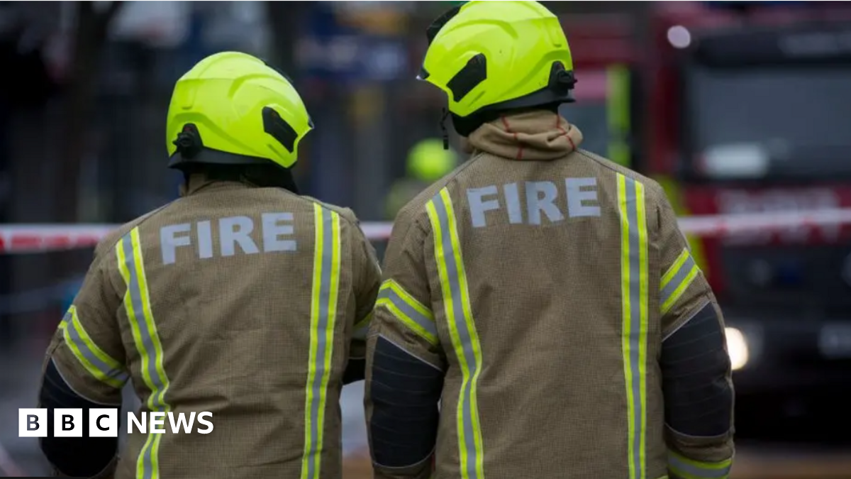 The back of two fire fighters. They are wearing a beige oversized jacket with the word "fire" printed in bold white lettering and wear illuminous yellow helmets