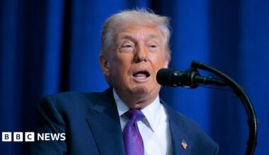 Donald Trump at a lectern with a blue suit, purple tie and US flag pin.