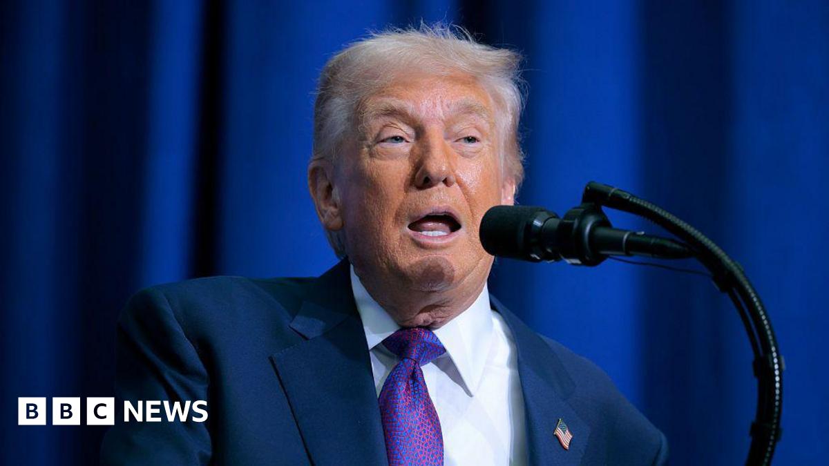 Donald Trump at a lectern with a blue suit, purple tie and US flag pin.