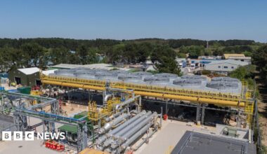 The United Downs power plant in Cornwall is shown, there are various silver pipes surrounding a central plant that has a series of large fan type structures surrounded by yellow pipes. There are trees in the background and the sky is blue.