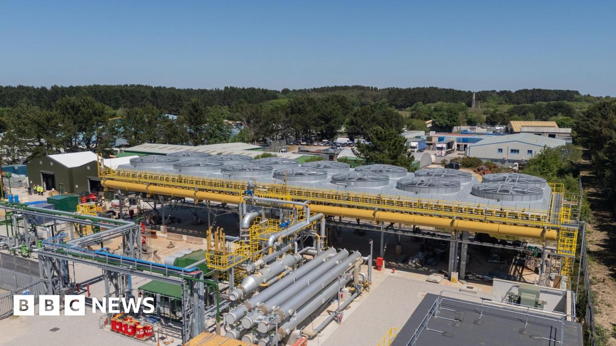 The United Downs power plant in Cornwall is shown, there are various silver pipes surrounding a central plant that has a series of large fan type structures surrounded by yellow pipes. There are trees in the background and the sky is blue.