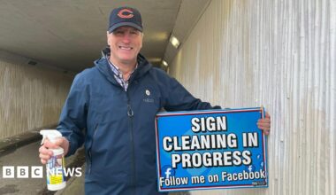 Photograph of John, who is looking into the camera and smiling. He is wearing a dark blue raincoat, with a purple checked shirt underneath. He is also wearing a dark blue cap with an orange letter C on it. He is holding a blue sign, that reads in white letters 'Sign Cleaning in Progress, follow me on Facebook', and in the other hand has a white spray bottle. He is standing in the underpass, which has cream coloured, corrugated walls which have large patches of white paint where he has begun to paint over the graffiti.
