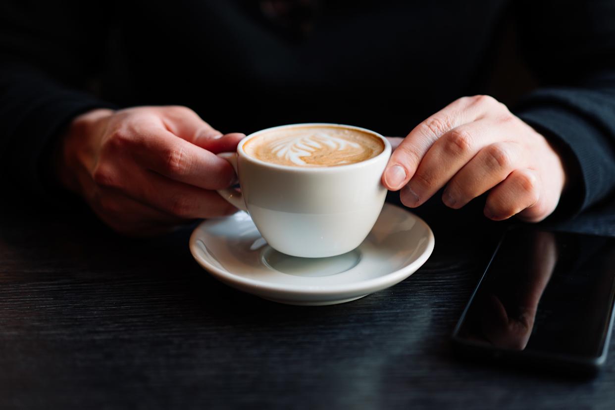 Hands holding a cappuccino with latte art on a saucer on a wooden table, suggesting a comfortable café moment.