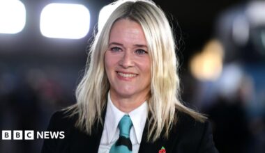 Edith Bowman is wearing a black suit jacket, a white shirt and a blue tie. She is smiling, with blurred bright lights behind her.