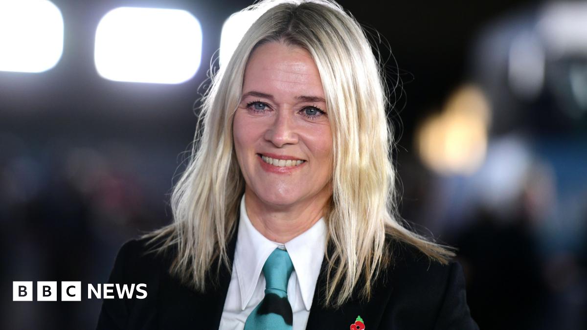 Edith Bowman is wearing a black suit jacket, a white shirt and a blue tie. She is smiling, with blurred bright lights behind her.