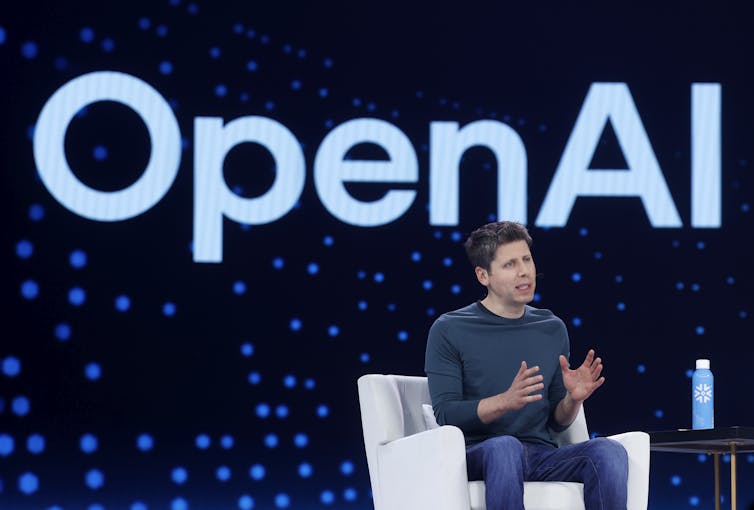 A man speaks while sitting in a chair in front of a wall emblazoned with OpenAI.