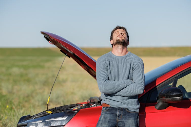 man looking despairing beside his car with its bonnet up.