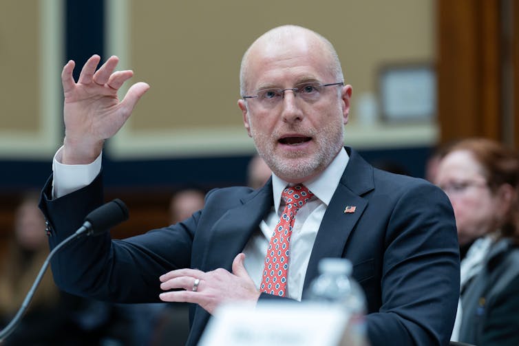 A man in suit in tie speaks in front of a microphone.