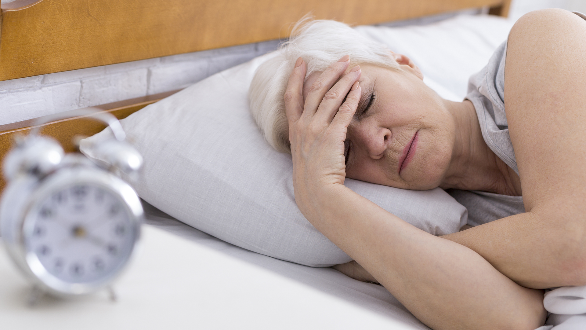 An older woman lies in bed with her head in her hands, struggling to sleep. At the front of the image is an out of focus alarm clock