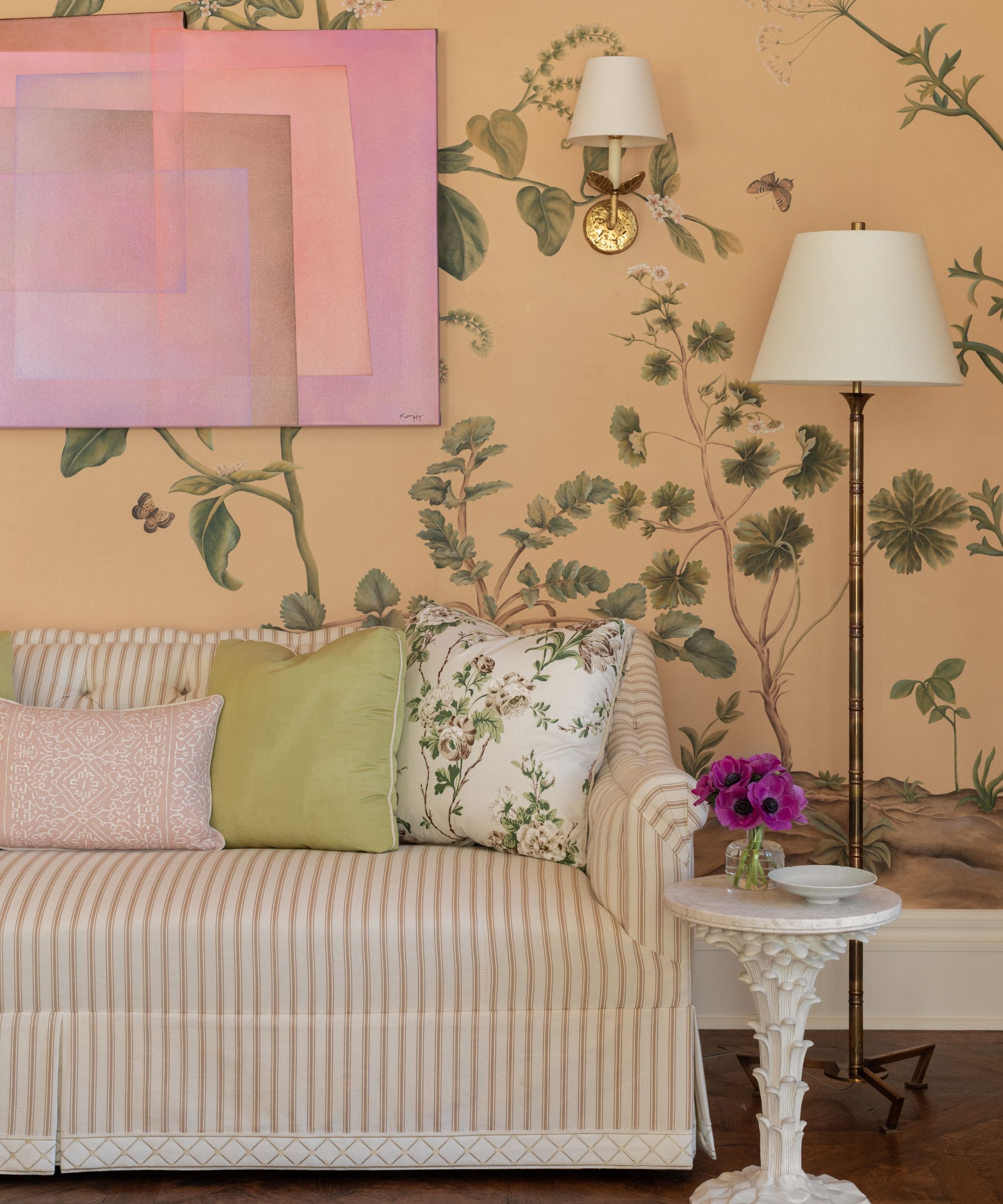 A living space with a botanical-scene wallpaper in a peach color, a light pink striped sofa, a piece of pink abstract artwork on the wall, and green and pink cushions of the sofa.