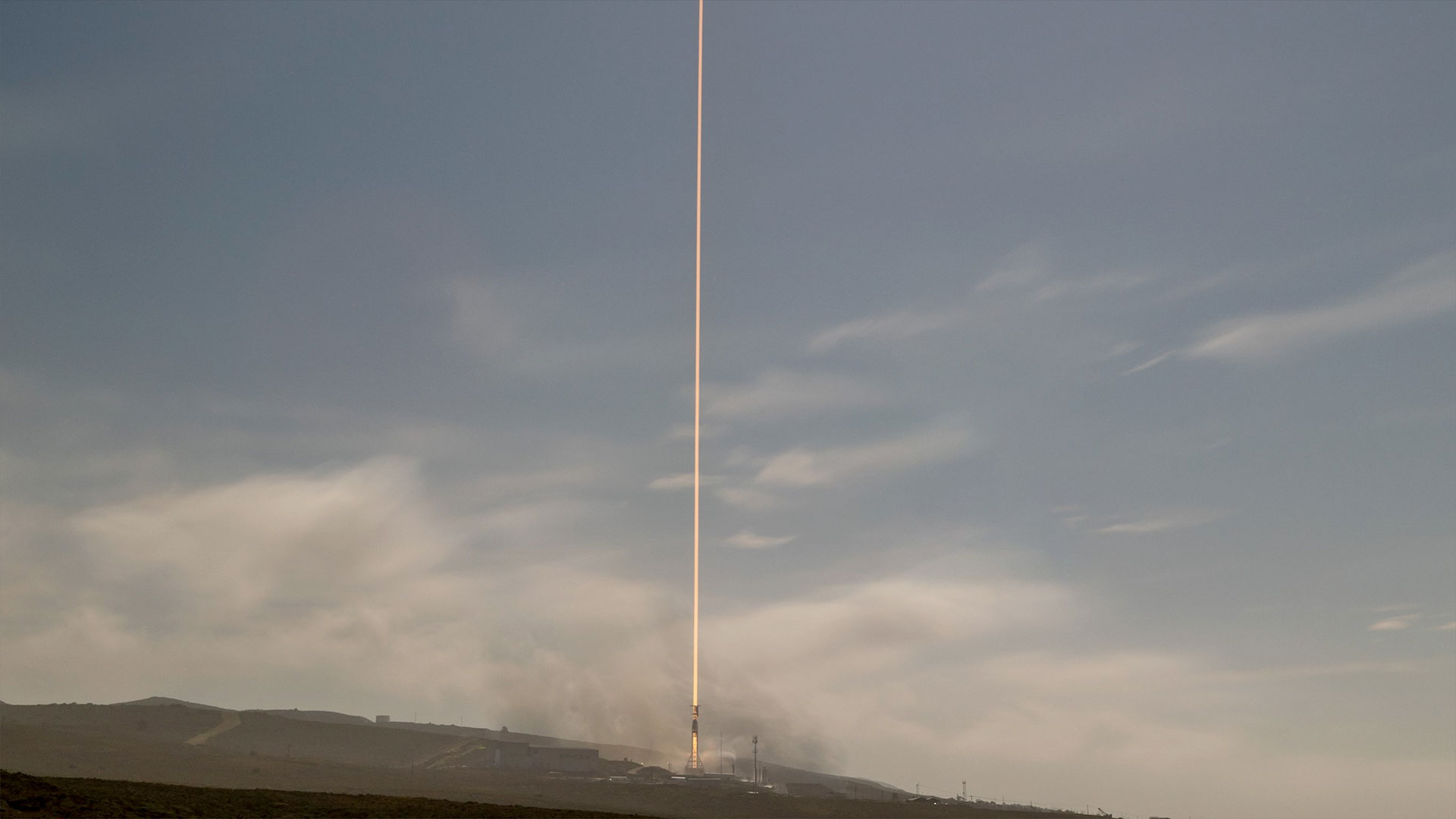 A daytime time lapse exposure of a rocket launch lifting off into partially cloudy sky.