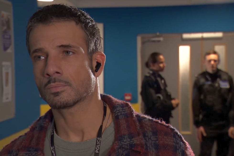 hollyoaks dodger savage in the police station corridor with an earpod in, and four uniformed officers in the background