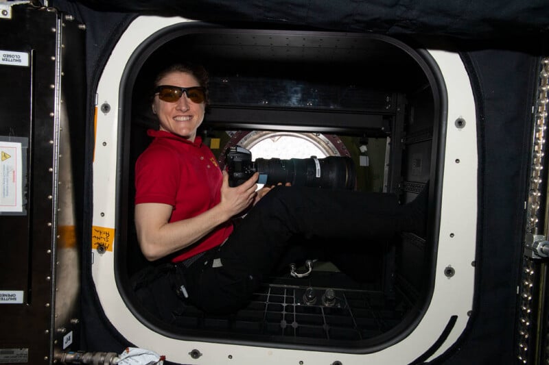 A person in a red shirt and sunglasses smiles while holding a camera, sitting inside a small hatch on what appears to be a space station, with equipment and a round window in the background.